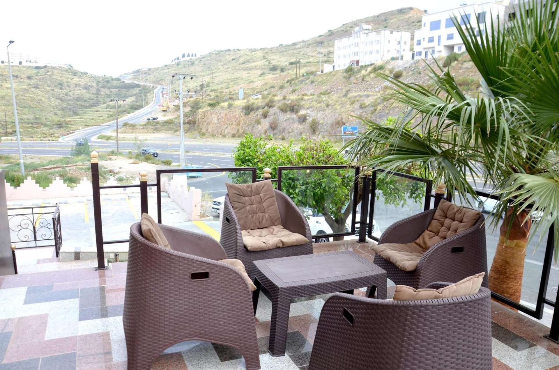 Balcony with rattan chairs, cushions and coffee table overlooking palm trees, hillside buildings and a winding road at فندق فربيون ابها - Ferbion Hotel Abha in Abha
